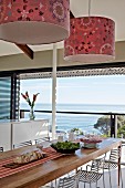 Pendant lamps with patterned, fabric lampshades above dining table and metal chairs next to panoramic window with sea view