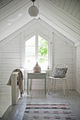 Table and chair in front of gable-end window in attic with white wooden cladding
