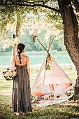 Frau in langem Sommerkleid mit Blüten-Korb vor romantischem Tipi-Zelt mit Picknick