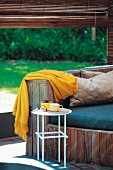 Wooden bench with seat cushions and yellow blanket in front of rolled up bamboo blind