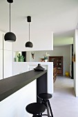 White fitted kitchen with black counter, matching pedant lamps, bar stools and doorway leading into living area