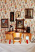 View across bed to 50s-style desk and chair below collection of pictures on floral wallpaper