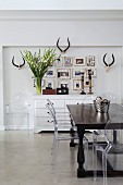 Colonial-style table, postmodern Ghost chairs, pictures and antlers above antique chest of drawers