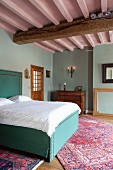 Double bed with turquoise upholstery and white bed linen in traditional bedroom with pink-painted wood-beamed ceiling and pastel turquoise walls