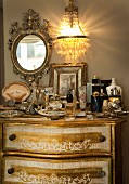 Chest of drawers with gilt details and jumble of ornaments on top below crystal sconce lamp and mirror in ornate gilt frame