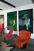 Retro orange leather armchair next to transparent shell chair in front of large modern artworks