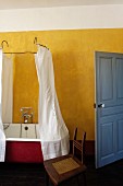 Vintage bathtub with shower curtain hanging from metal frame in rustic bathroom with yellow wall and blue door