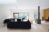 Lounge area with black sofas and free-standing wood-burner in minimalist interior; man sitting on window seat