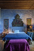 Rustic bedroom with religious painting on blue-painted wall, double bed with blue bedspread and purple blanket on trunk at foot of bed