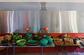 Green ceramic crockery on metal table and stacked basket trays on side table below stainless steel cladding on wall