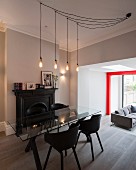 Light-bulb pendant lamp above glass table and black shell chairs in open-plan interior with red steel structure in background