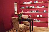 Dark wooden table and two rattan chairs in front of narrow DIY shelves on claret-red wall