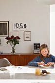 Girl eating at marble table in front of vases of roses on sideboard below framed pictures