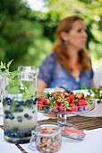 Refreshing drink with blueberries in carafe and fresh strawberries in glass dish on garden table
