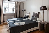 Double bed with printed, charcoal grey bedspread flanked by tall lamps on modern bedside tables against white, wood-clad wall