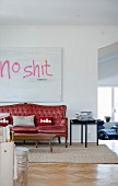 Red, antique sofa, rustic wooden table and delicate side table below artwork with motto on wall of open-plan living area