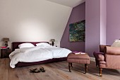 Doppelbett mit weisser Bettwäsche, davor gemütlicher Sessel mit passendem Fussschemel, vor violett getönter Wand im Dachgeschoss