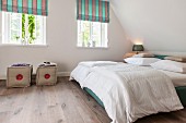 Schlafzimmer in modern ländlichem Stil, Doppelbett mit weisser Bettwäsche unter Dachschräge, seitlich Fenster mit gestreiften Raffrollos und Polsterhocker