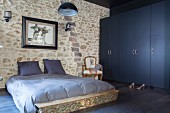Grey-blue bed linen on double bed against stone wall next to dark blue wardrobes