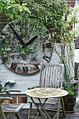 Weathered table and wooden chairs on terrace in front of vintage station clock face on climber-covered brick wall