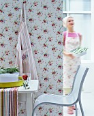 Modern chair and table in front of partition with floral wallpaper; woman wearing apron and headscarf in background