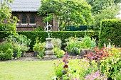 Flowering perennials and armillary sundial on stone plinth in summery garden in front of hedge and brick façade