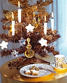 Small, gold artificial Christmas tree decorated with lit candles next to elegant coffee cup and plate on shiny side table
