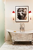 Free-standing, silver bathtub with vintage-style wall-mounted taps below red sconce lamps and feminine photograph