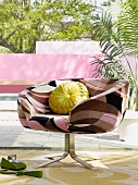 Retro swivel chair with patterned cover and yellow scatter cushion on terrace in front of pool and garden wall painted pink