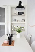 Vase of ranunculus and beaker of black drinking straws on white dining table in front of open-fronted shelves of crockery