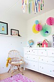 Mädchenzimmer mit Korbstuhl, weißem Schubladenschrank und bunten Papier-Pompons