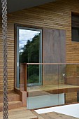 Fassadenausschnitt mit Holzverkleidung, Glastür und Metallplatte, davor Glasbrüstung und Treppenstufen