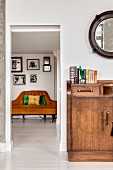 Retro Holzkommode mit Büchern, darüber runder Wandspiegel und Blick auf Sofa in Nebenraum