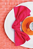 Geknotete Stoffserviette und orangefarbene Tasse auf weißem Teller