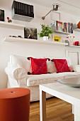 Red cushions on white loose-covered sofa below wall-mounted shelves
