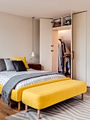 Bed with yellow upholstered frame next to open fitted wardrobe under sloping ceiling