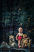 Macabre arrangement of dolls, dolls' furniture and feathers arranged against dark background