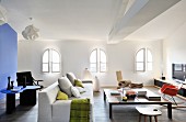Helles Sofa, Couchtish, Designerstühle und Lowboard im Loungebereich mit Rundbogenfenstern; blauer Raumteiler und Designertisch