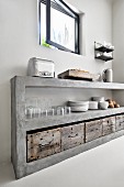 Crockery and glasses stacked on concrete kitchen shelves above rustic wooden crates below open window