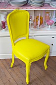 Antique upholstered dining chair painted yellow in front of pink glass vessels and stacked white plates in white dresser