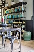 Farbige Sodaflaschensammlung und Keramikgefässe in offenem Regal hinter rustikalem Tisch und Stühlen