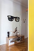 Yellow door frame and view of mirror shaped like sunglasses with black frame next to white coat rack in hall