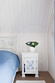 Nostalgisches, weißes Nachtkästchen neben Bett mit Vintage Betthaupt unter holzverkleideter Dachschräge