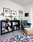 Black bookcase on castors next to green leather armchair in corner and footstool on colourful rug