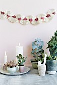 Arrangement of plants and lit white candles below garland on wall