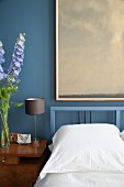 Schlafzimmer mit blauer Wand, gerahmtem Bild und Blumen auf Nachtkästchen