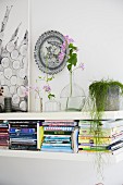 Several stacks of books on wall-mounted shelf and delicate flower arrangements below decorative wall plate