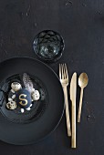 Place setting with gold cutlery, black plate and arrangement of eggs on glass plate
