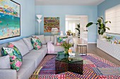 Living room with pastel blue walls, gray corner couch and glass coffee table on colorfully patterned carpet