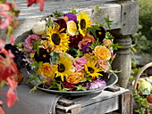 Wreath of sunflowers, roses and dahlias on wine box ajar against bench